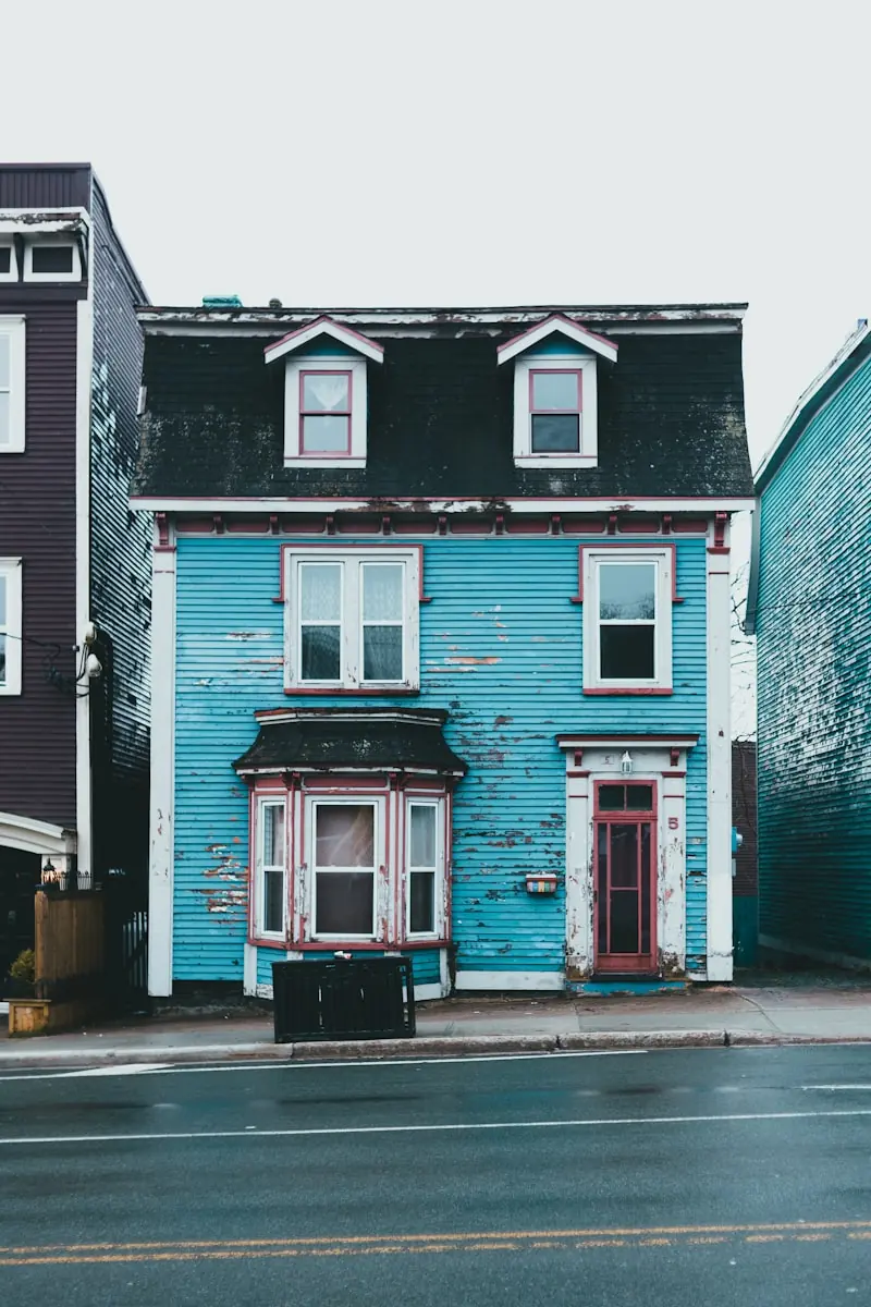 A blue clapboard house with two dormer windows and red trim between neighbouring houses